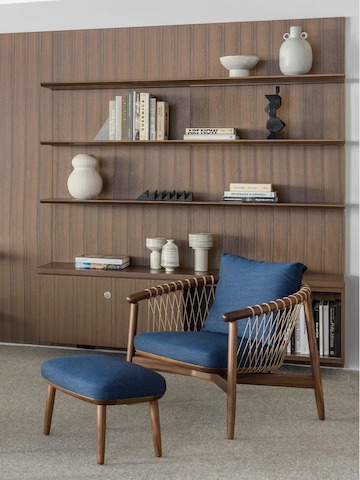 A Crosshatch Chair and Ottoman with dark blue fabric and a wood frame in a medium finish, positioned at an angle in front of bookshelves.