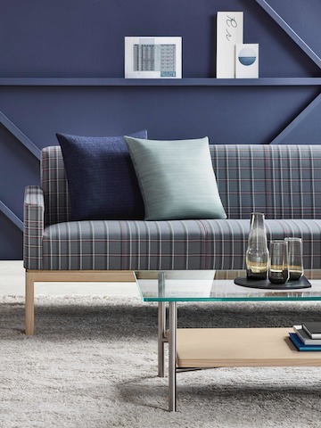 Partial view of a rectangular Layer coffee table with glass and wood shelves in front of a Wood Base Lounge Seating sofa with blue plaid fabric.