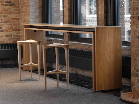 A rectangular Peer Table and a pair of complementary 2 by 3 Stools positioned against three windows in a brick-walled office space.