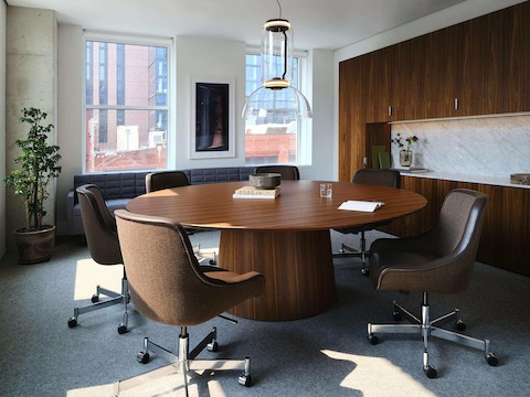 JD Meeting Table by DatesWeiser in Natural Quarter Cut Walnut with round top and tapered base shown at an angle surrounded by Bumper Chairs in front of a Geiger One workwall.