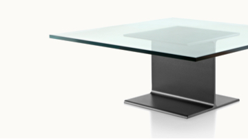 Partial angled view of a square I Beam coffee table with a glass top and black cast-aluminum pedestal.