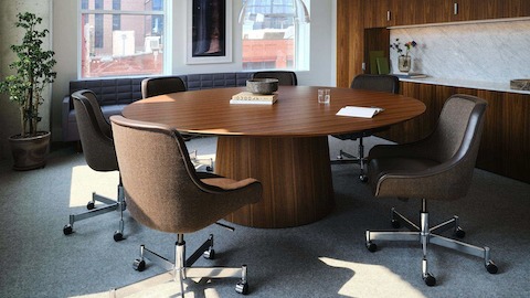 JD Meeting Table by DatesWeiser in Natural Quarter Cut Walnut with round top and tapered base shown at an angle surrounded by Bumper Chairs in front of a Geiger One workwall.