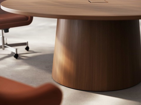 Detail of JD Meeting Table by DatesWeiser showing tapered round base in natural quarter cut walnut with two bumper chairs on a concrete floor.