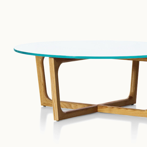 Partial view of a round coffee table with a glass top and a wood base in a medium finish. Select to go to the Loophole Tables product page.